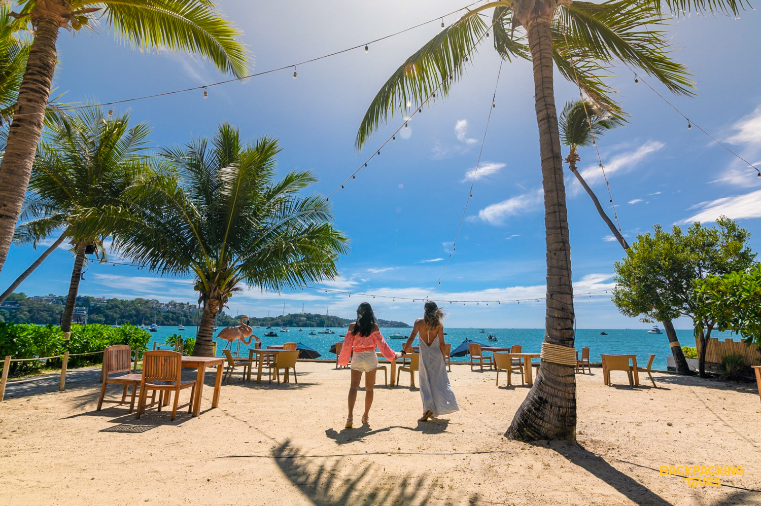 Friends stroll hand-in-hand on a sunny Phuket beach, past outdoor dining, towards the sparkling ocean.