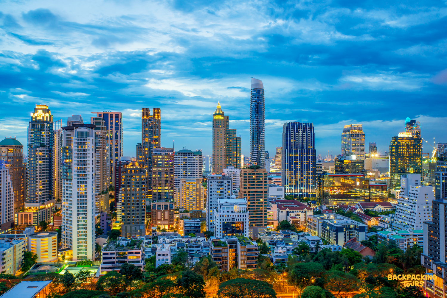 A vibrant Bangkok cityscape at dusk, featuring numerous illuminated skyscrapers under a dramatic cloudy sky.