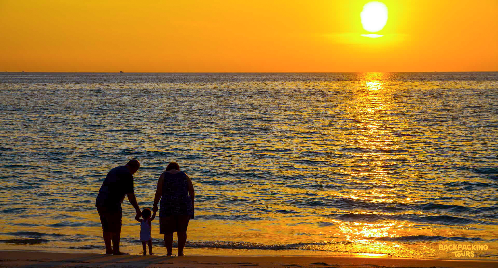 A family of three enjoys a beautiful sunset over the ocean on a beach in Phuket.