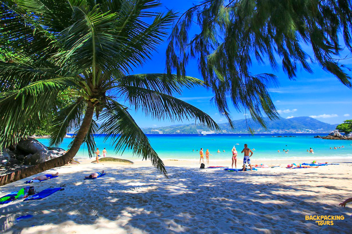 Tourists relax on a sunny white sand beach with turquoise water and palm trees in Phuket, Thailand.