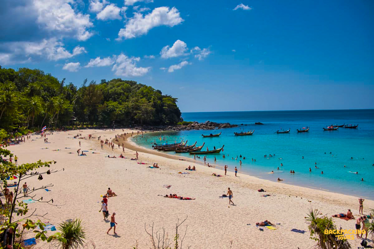 A bustling Phuket beach with sunbathers, swimmers, and traditional longtail boats in the clear water.