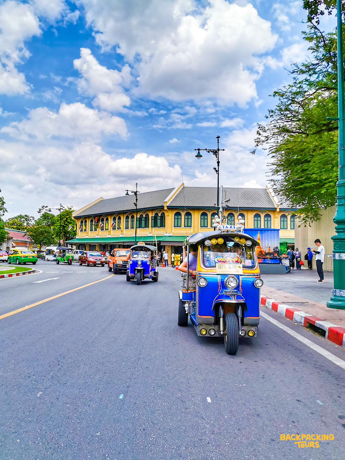 Blue tuk-tuks transport people on a bustling street in Bangkok with a large yellow building.