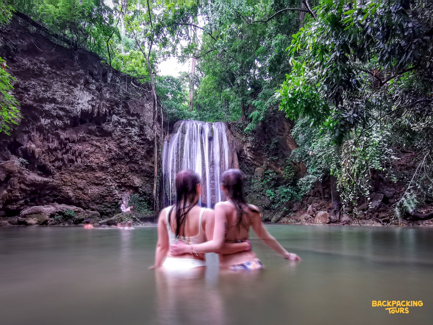 Two friends embrace in the refreshing emerald pools of Erawan Waterfalls, surrounded by lush jungle.