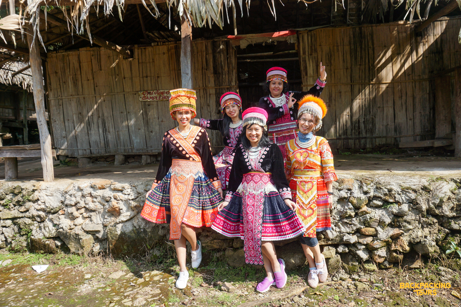 Six smiling women in vibrant traditional Hmong clothing pose outside a rustic village house in Chiang Mai.