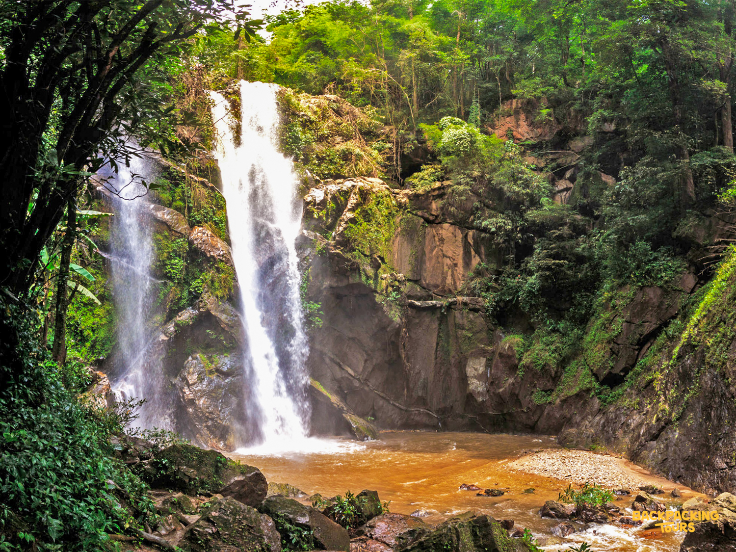 A powerful waterfall cascades into a brown pool, surrounded by lush green jungle in Chiang Mai.