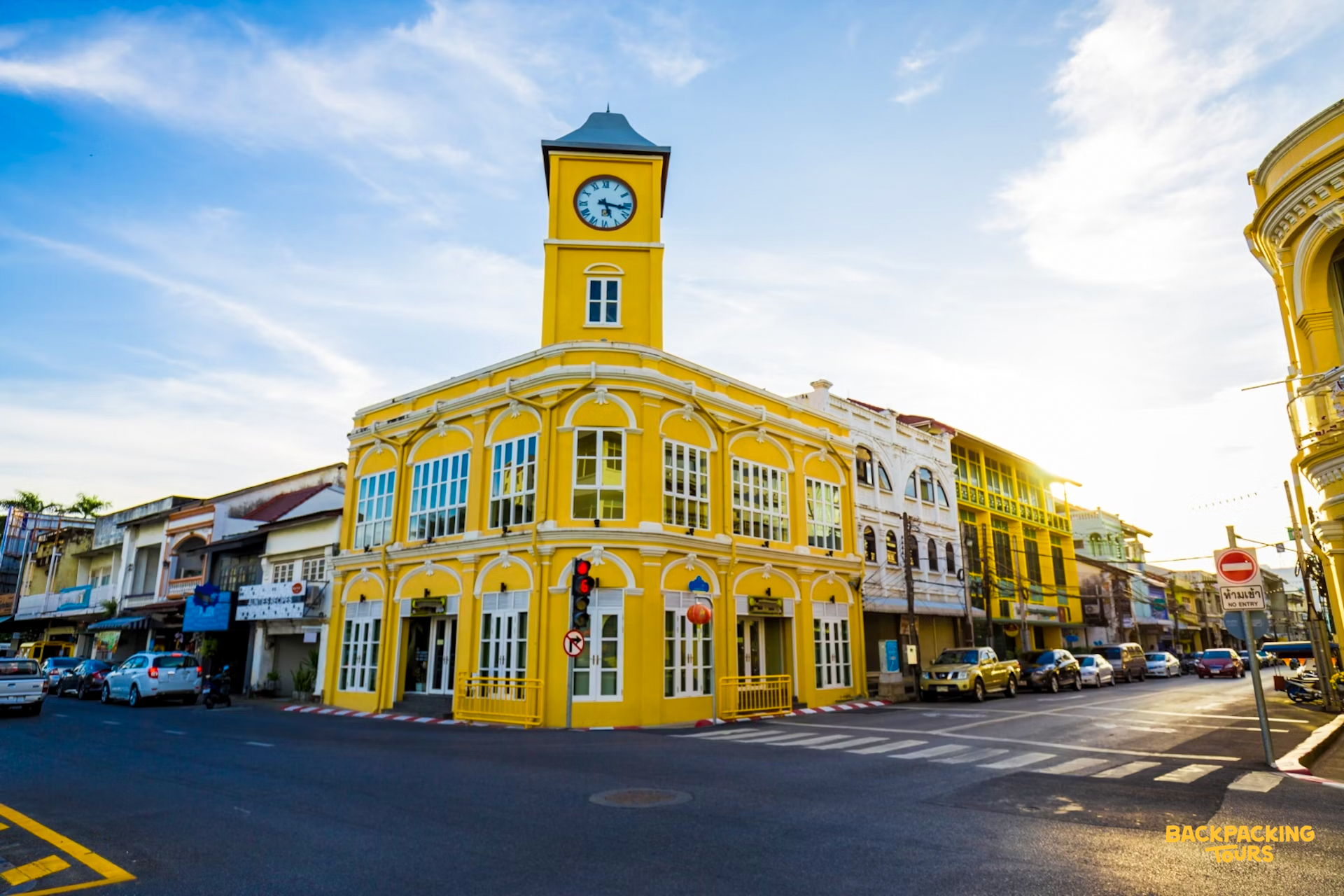 A vibrant yellow clock tower building stands on a sunny street corner in Phuket Old Town.
