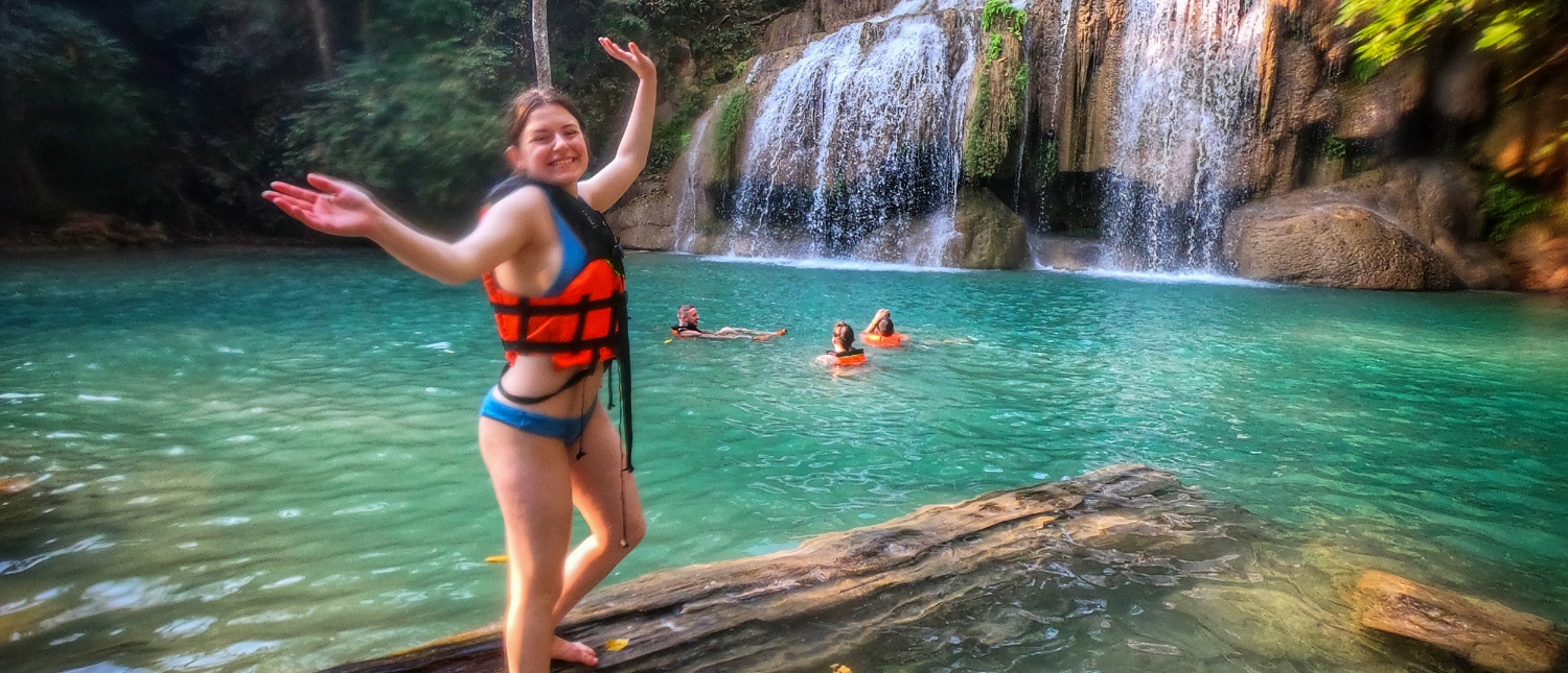 Joyful woman in a life vest on a log in emerald pools at Erawan Waterfalls, Thailand.