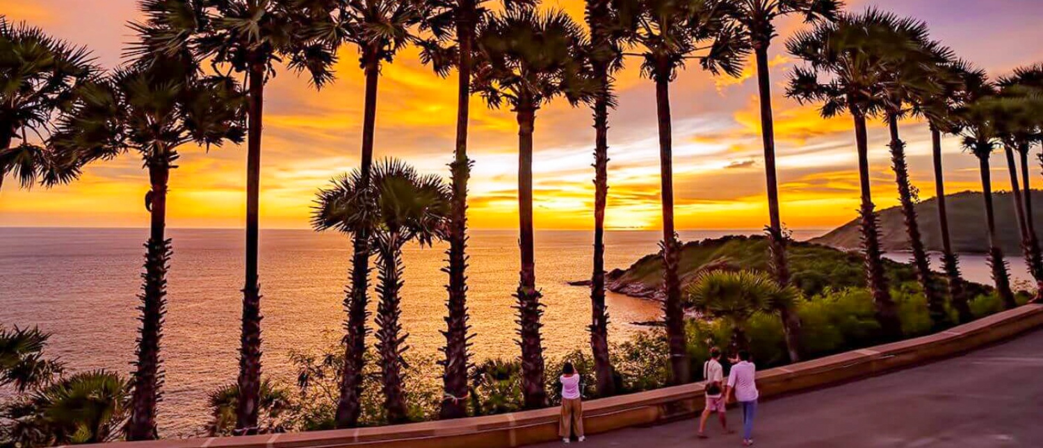 Tourists watch a vibrant sunset over the ocean, framed by tall palm trees in Phuket, Thailand.