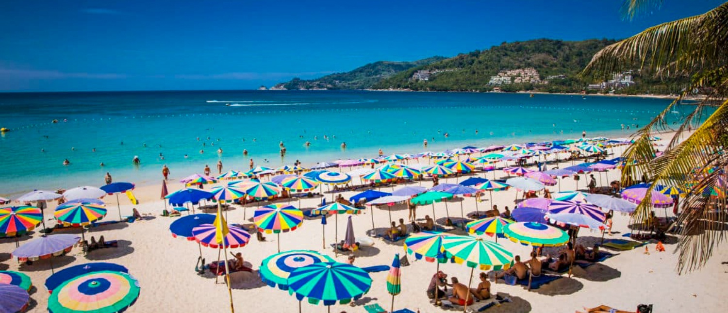 Colorful umbrellas line a beautiful tropical beach in Phuket with people swimming in turquoise water.