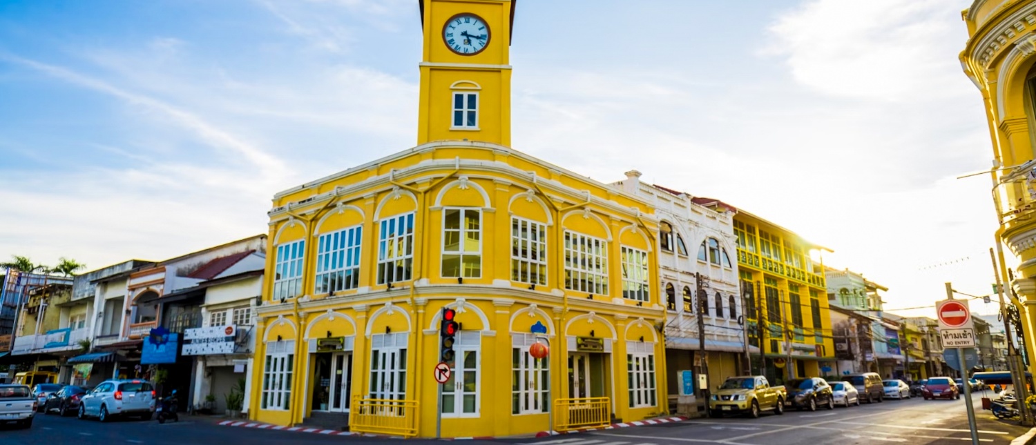A vibrant yellow clock tower building anchors a sunny street lined with colorful architecture in Phuket.