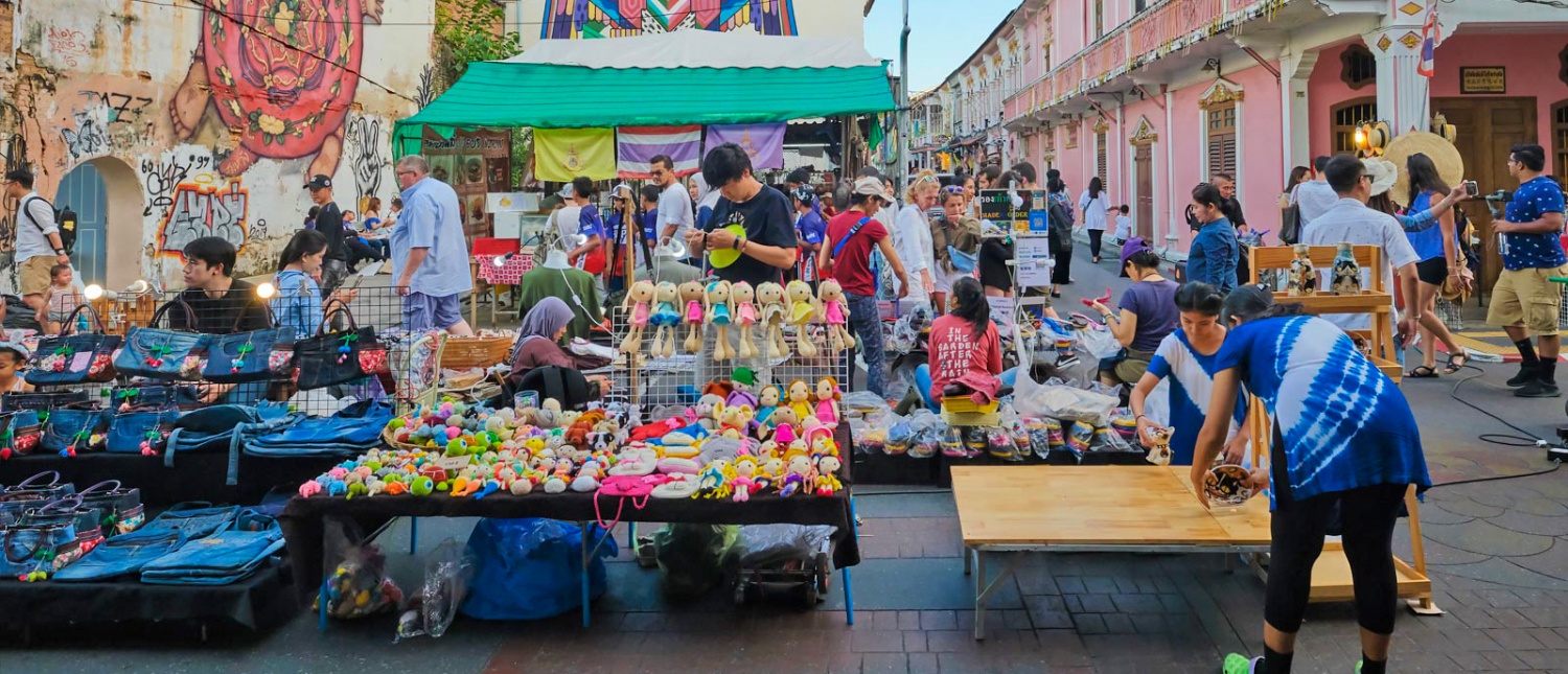 A bustling street market in Phuket Old Town with people browsing colorful stalls selling various goods.