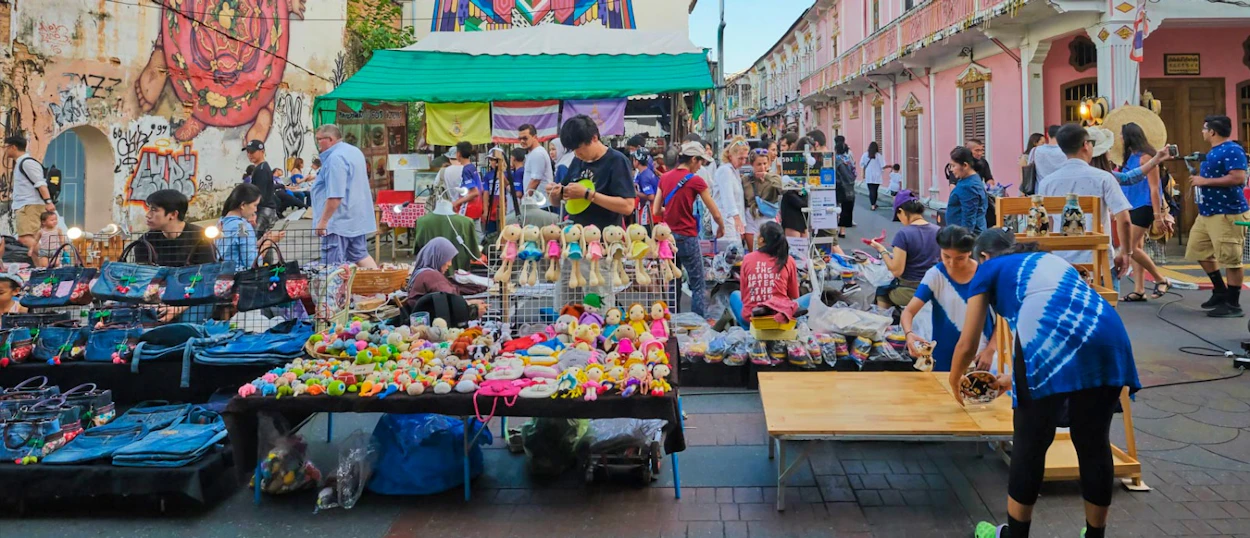 A bustling street market in Phuket Old Town with people browsing colorful stalls selling various goods.