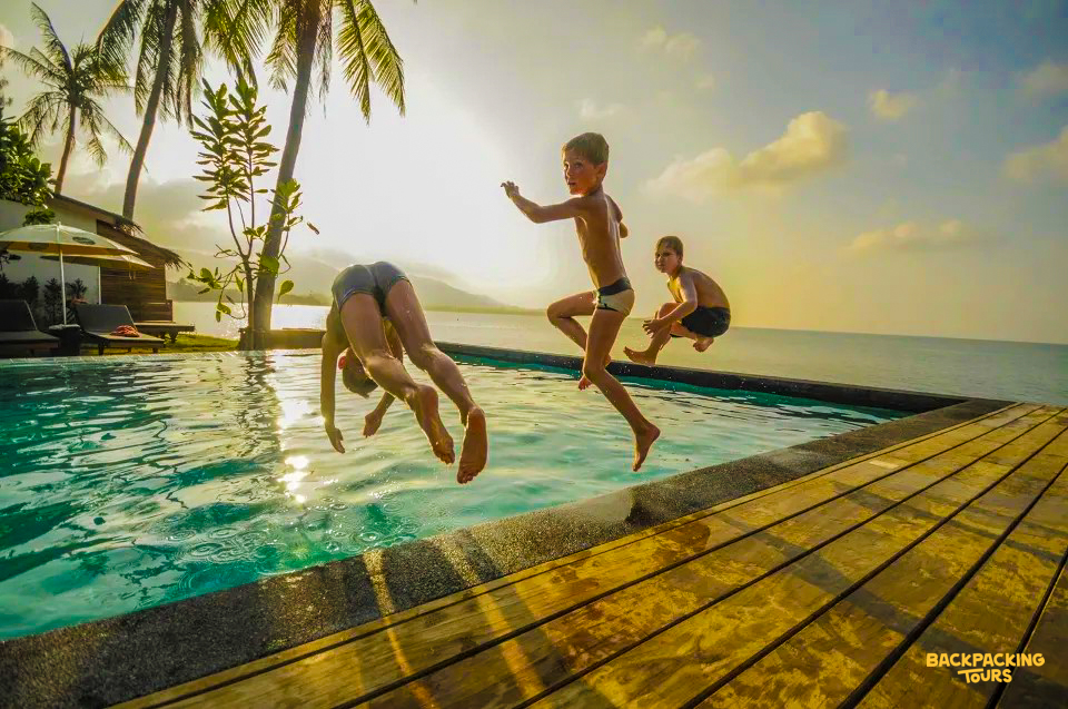 Three boys happily jump into an infinity pool overlooking the ocean at sunset in Koh Samui.