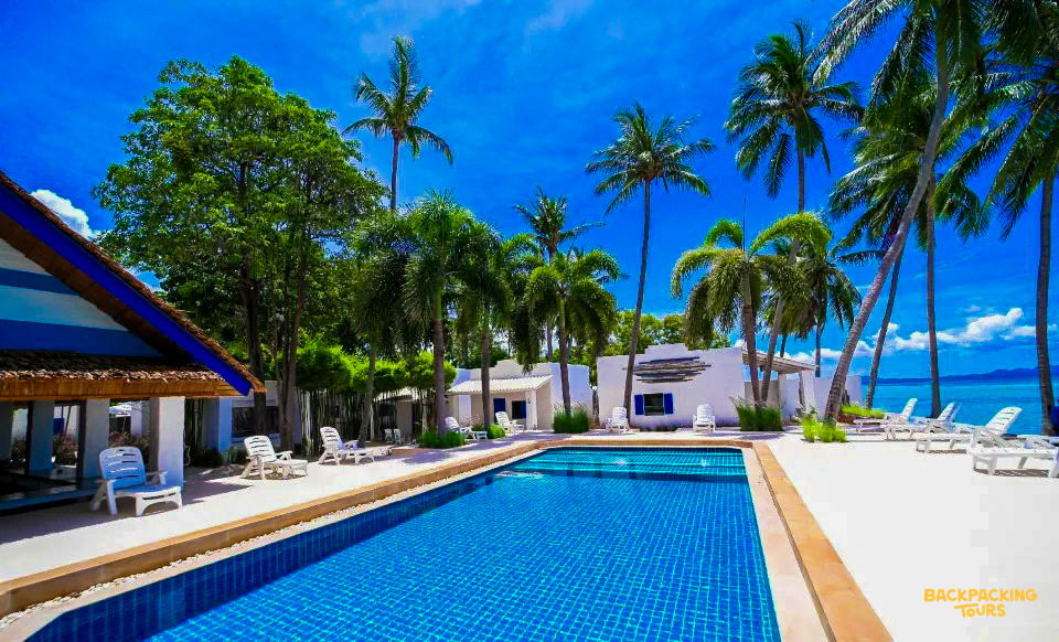 Tropical resort on Koh Phangan featuring a sparkling blue pool, white buildings, and swaying palm trees.