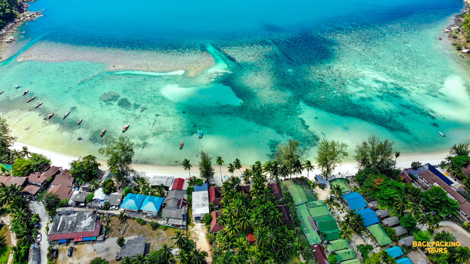 Vibrant turquoise bay with small boats, white sand, and colorful beachfront buildings on Koh Phangan.