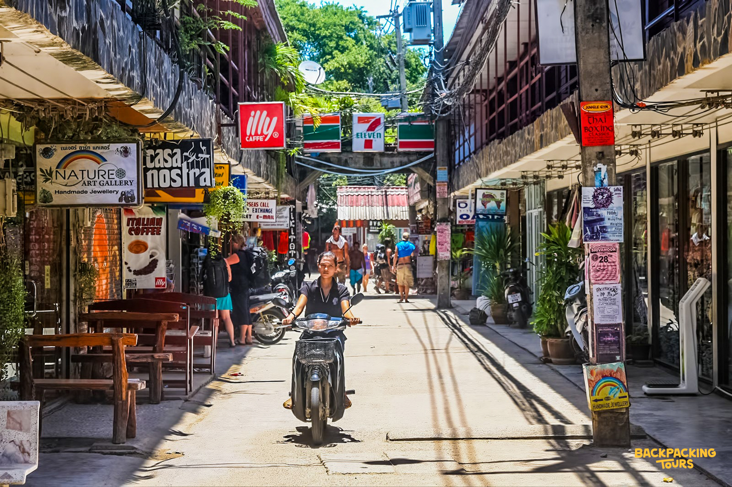A person rides a scooter down a lively, narrow street lined with shops in Koh Phangan.