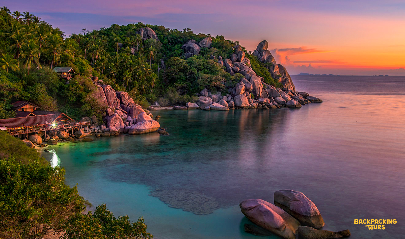 Vibrant sunset over a serene bay on Koh Phangan, with lush tropical hills and clear waters.