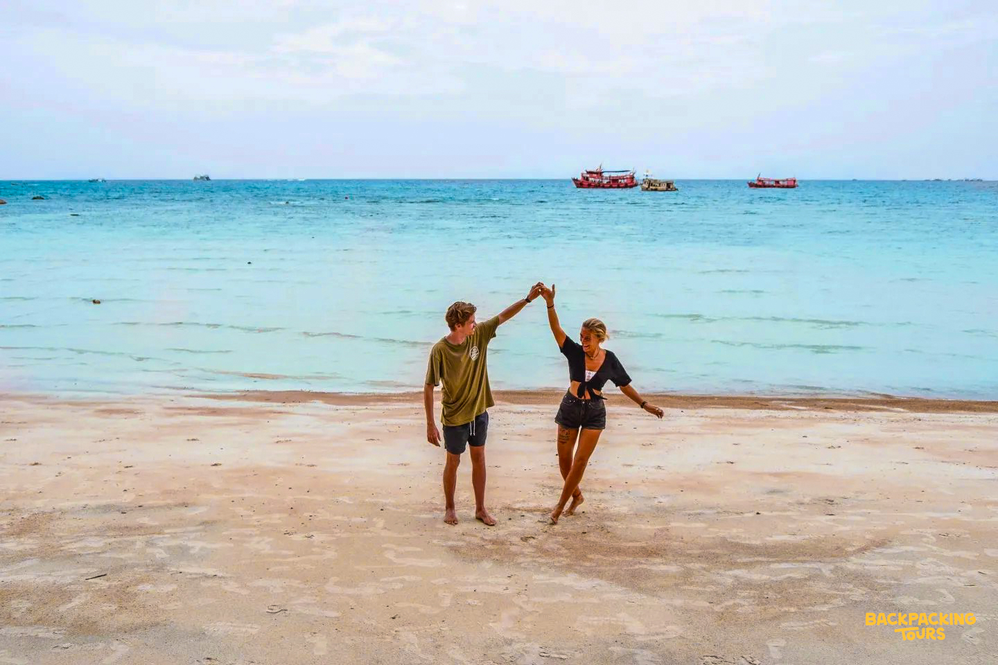 A happy couple dances on Sairee Beach, Koh Tao, with turquoise water and boats in the background.