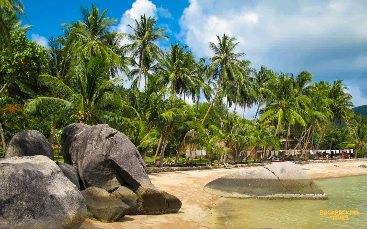 Lush palm trees and large rocks adorn the sandy Sairee Beach on a sunny day in Koh Tao.