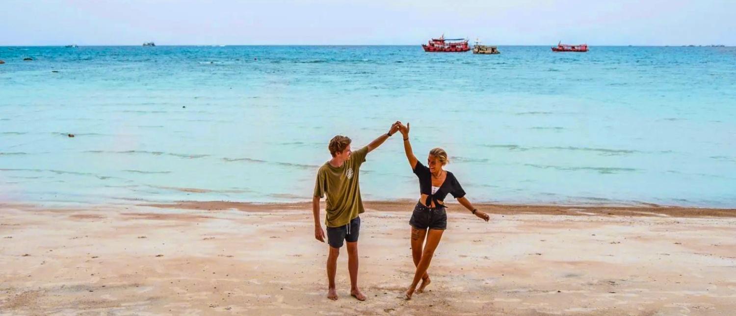 A happy couple dances playfully on Sairee Beach, Koh Tao, with blue ocean and boats.