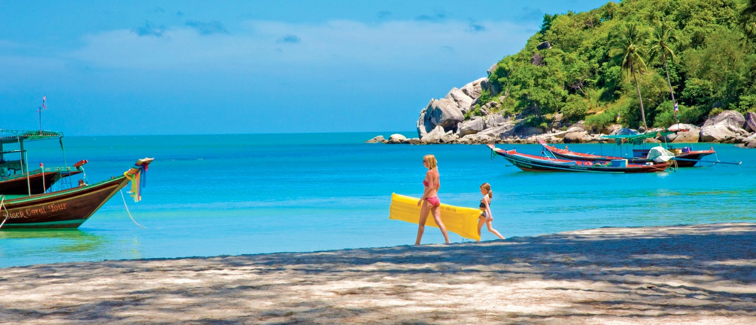 Mother and child walk on a sunny Koh Phangan beach with longtail boats in blue water.
