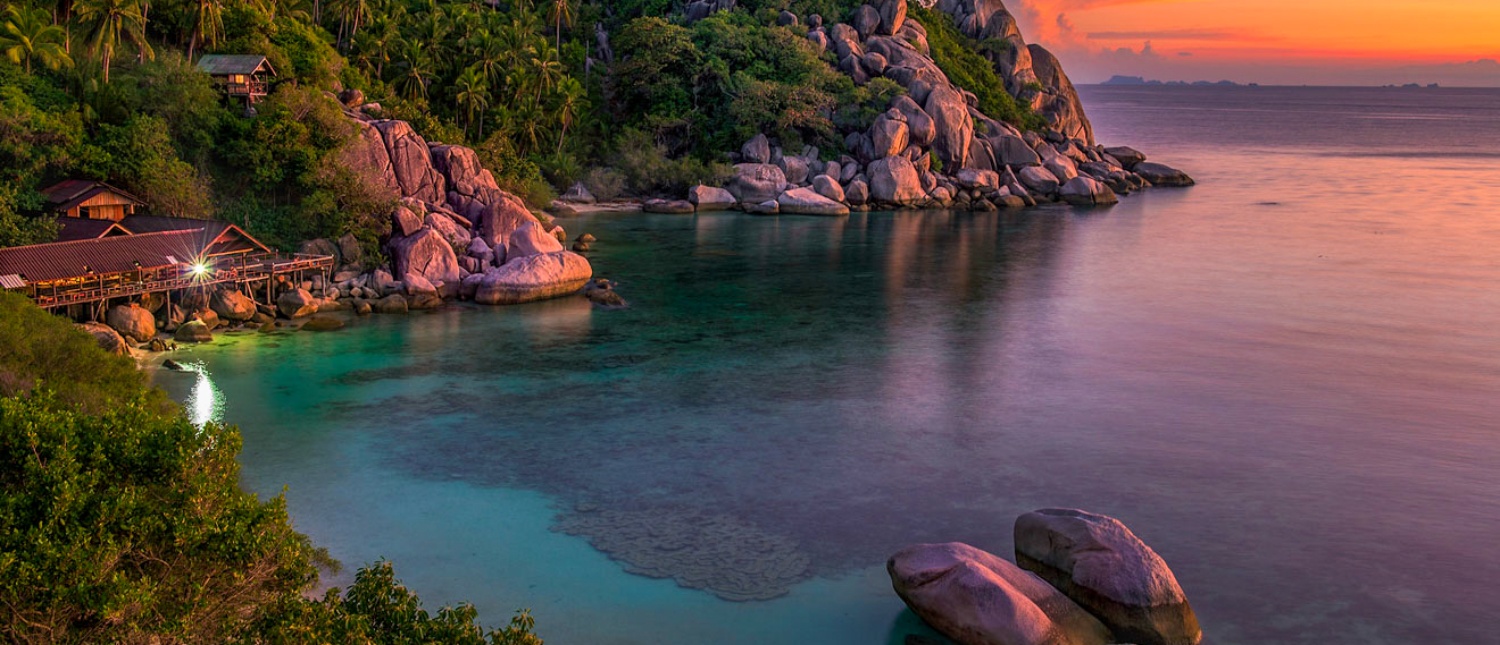 Tranquil tropical bay on Koh Phangan at sunset, with clear water, rocky shores, and a lit building.