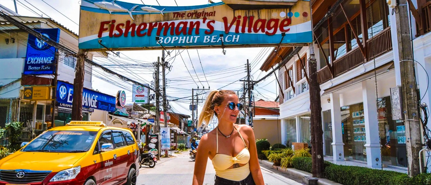 A smiling woman in sunglasses walks through the vibrant Fisherman's Village, Bophut Beach, Koh Samui.