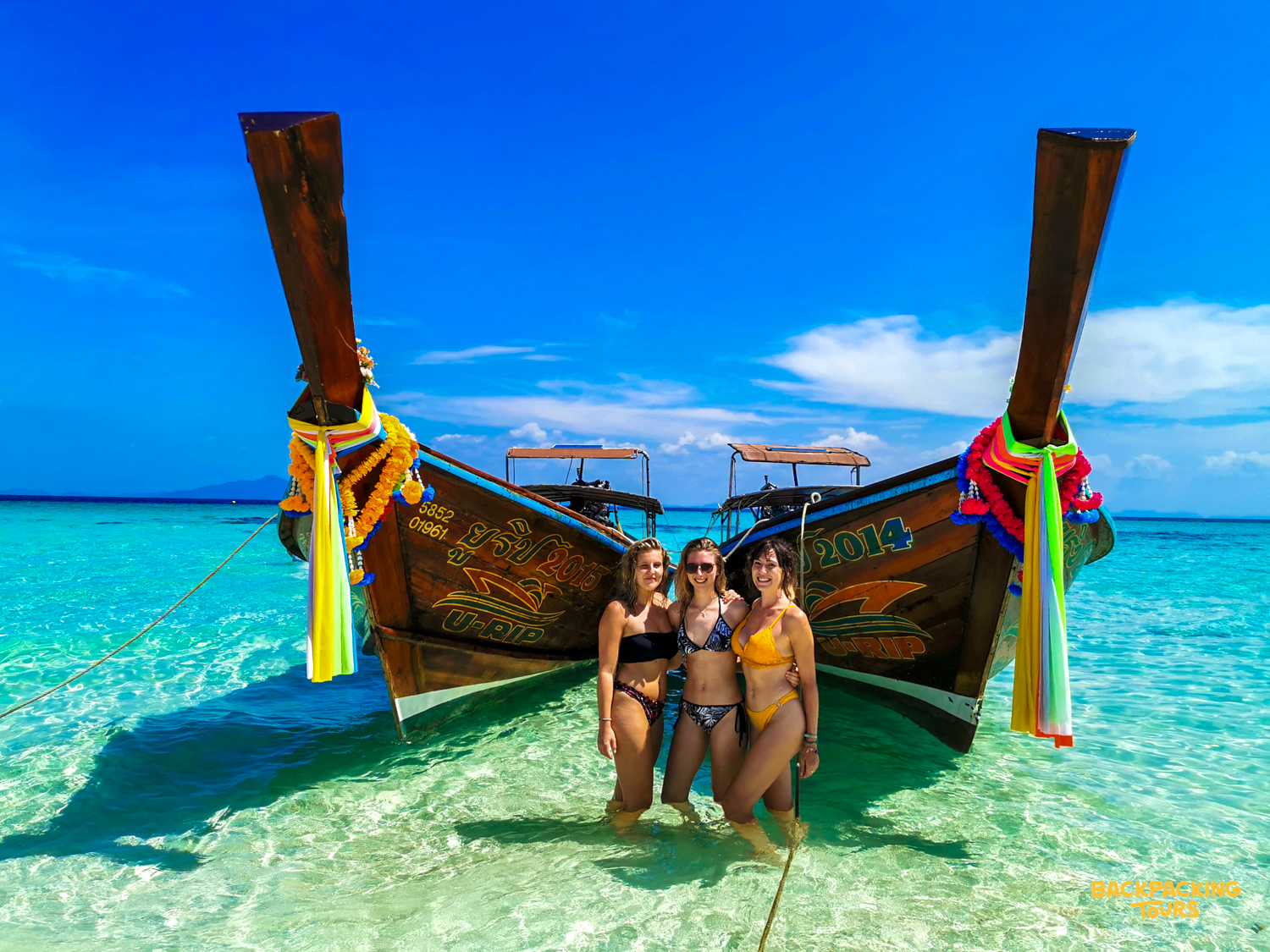 Three friends enjoy island time, posing in clear water with longtail boats on Phi Phi Islands.