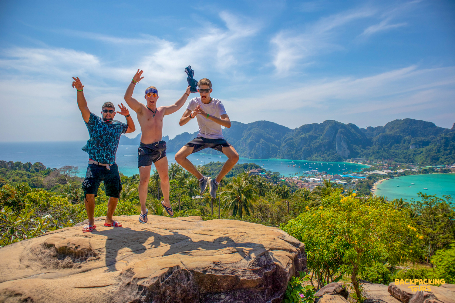 Three happy men jump for joy on a viewpoint overlooking the stunning Phi Phi Islands bay.