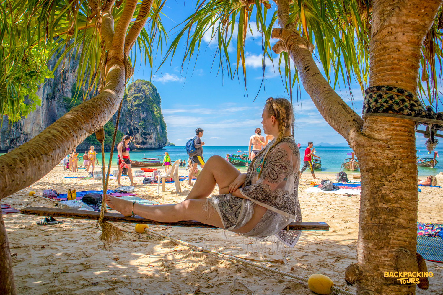 A woman relaxes on a swing under palm trees, enjoying the vibrant beach scene in Krabi.