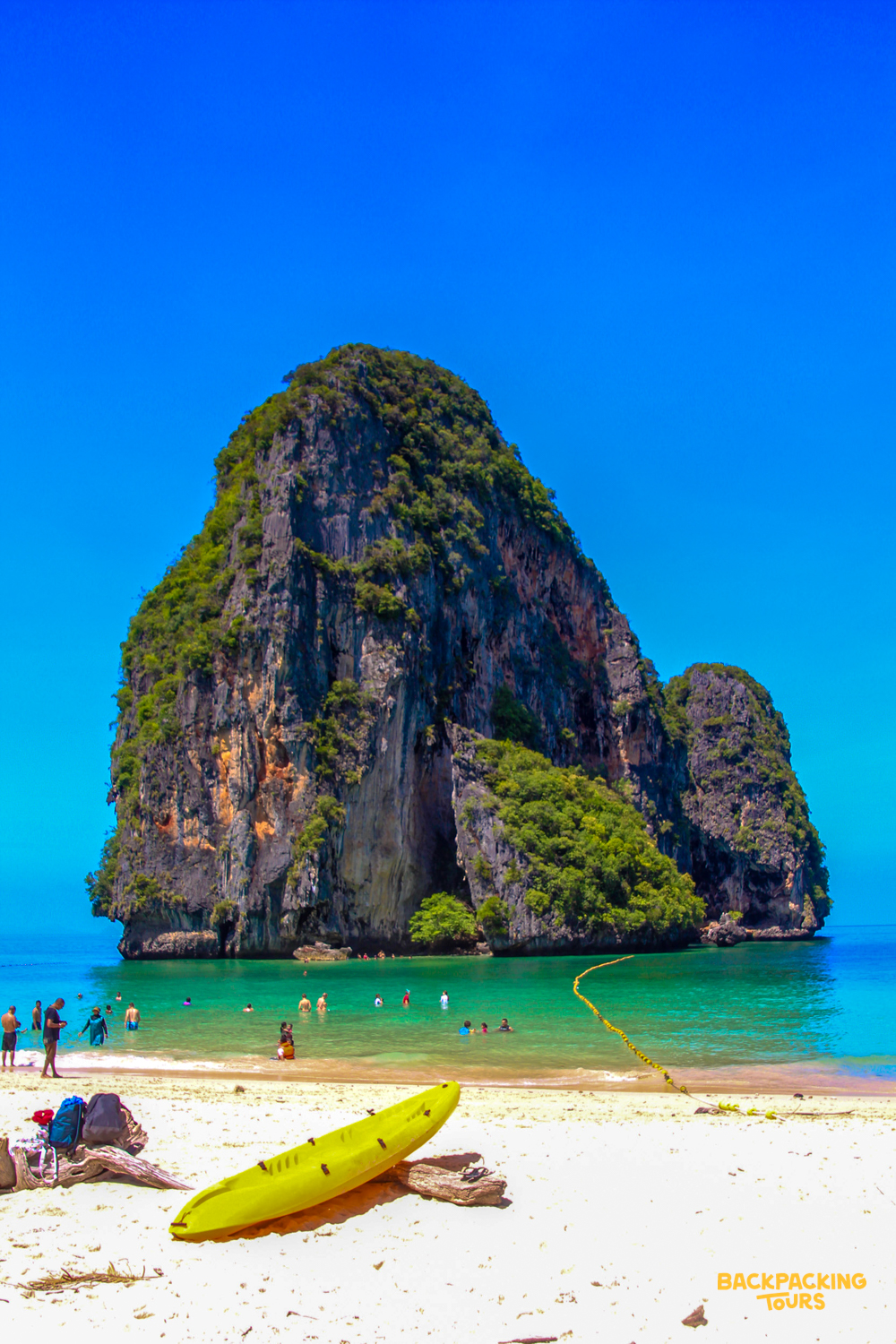 People swim and relax on a white sand beach with a towering limestone island in Krabi, Thailand.