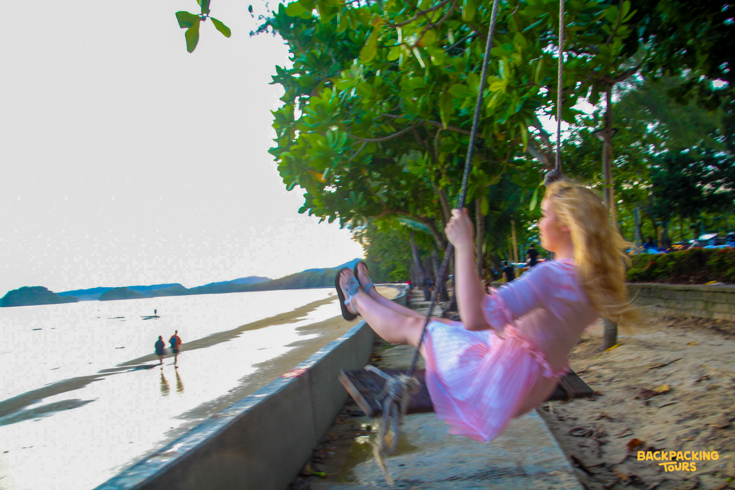 A young woman in a pink dress joyfully swings on a beach swing by the sea in Krabi.