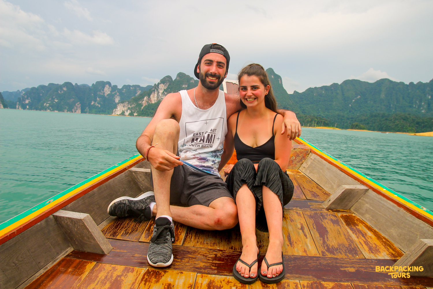 A happy couple smiles on a wooden boat, surrounded by towering karsts on Cheow Lan Lake.