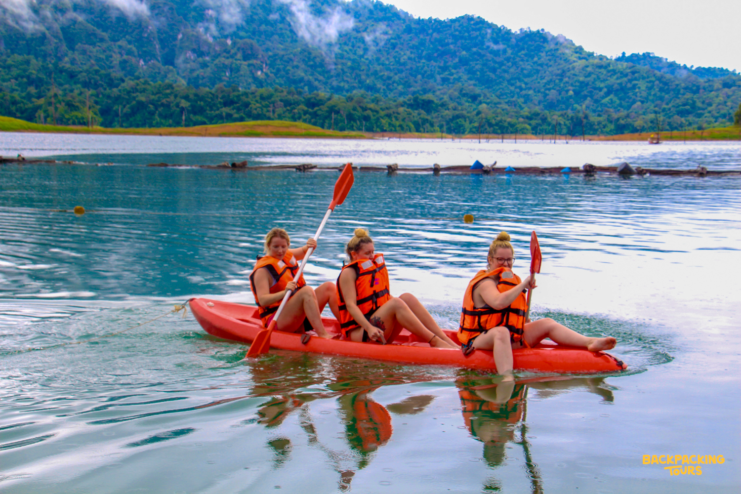 Three women paddle a red kayak on Cheow Lan Lake, surrounded by lush mountains in Khao Sok National Park.
