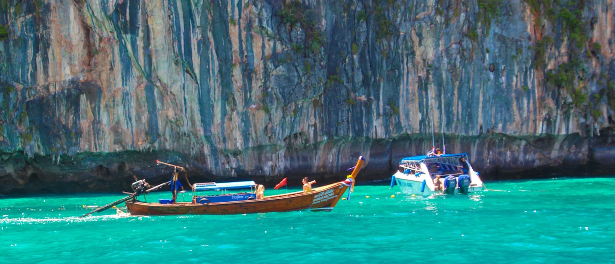 A traditional longtail boat and a speedboat navigate turquoise waters past dramatic limestone cliffs in Phuket.