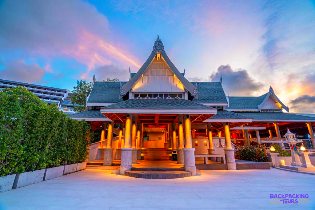 An elegant Thai-style resort building in Phuket, beautifully illuminated by warm lights under a vibrant sunset sky.