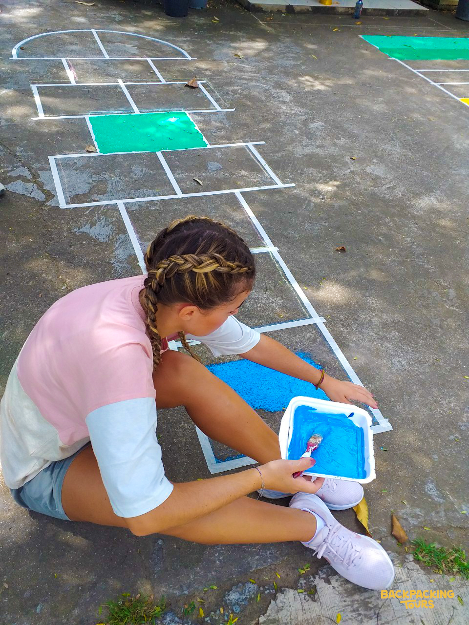 A volunteer paints a hopscotch court with bright blue and green colors on a concrete surface.