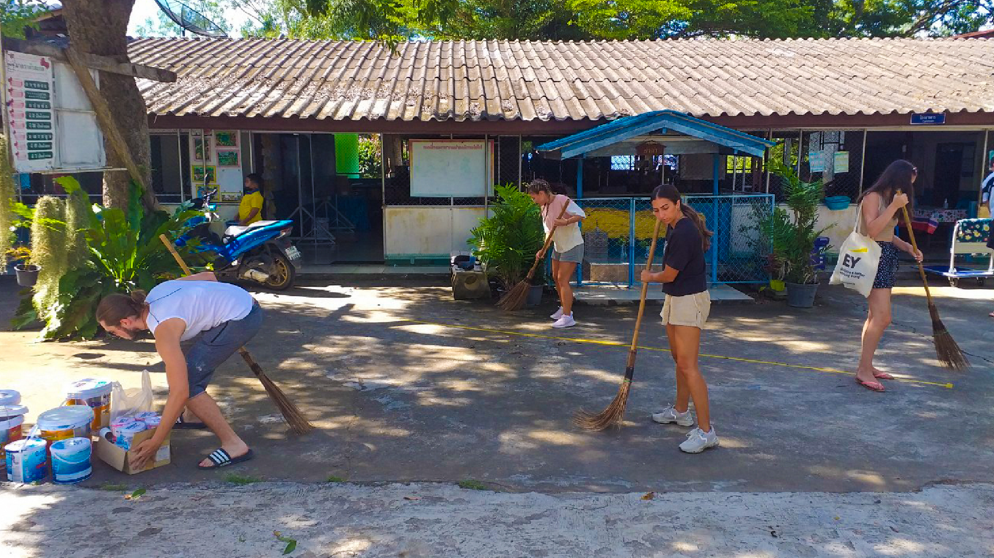 Four volunteers sweep a paved area and organize construction supplies outside a building in Thailand.