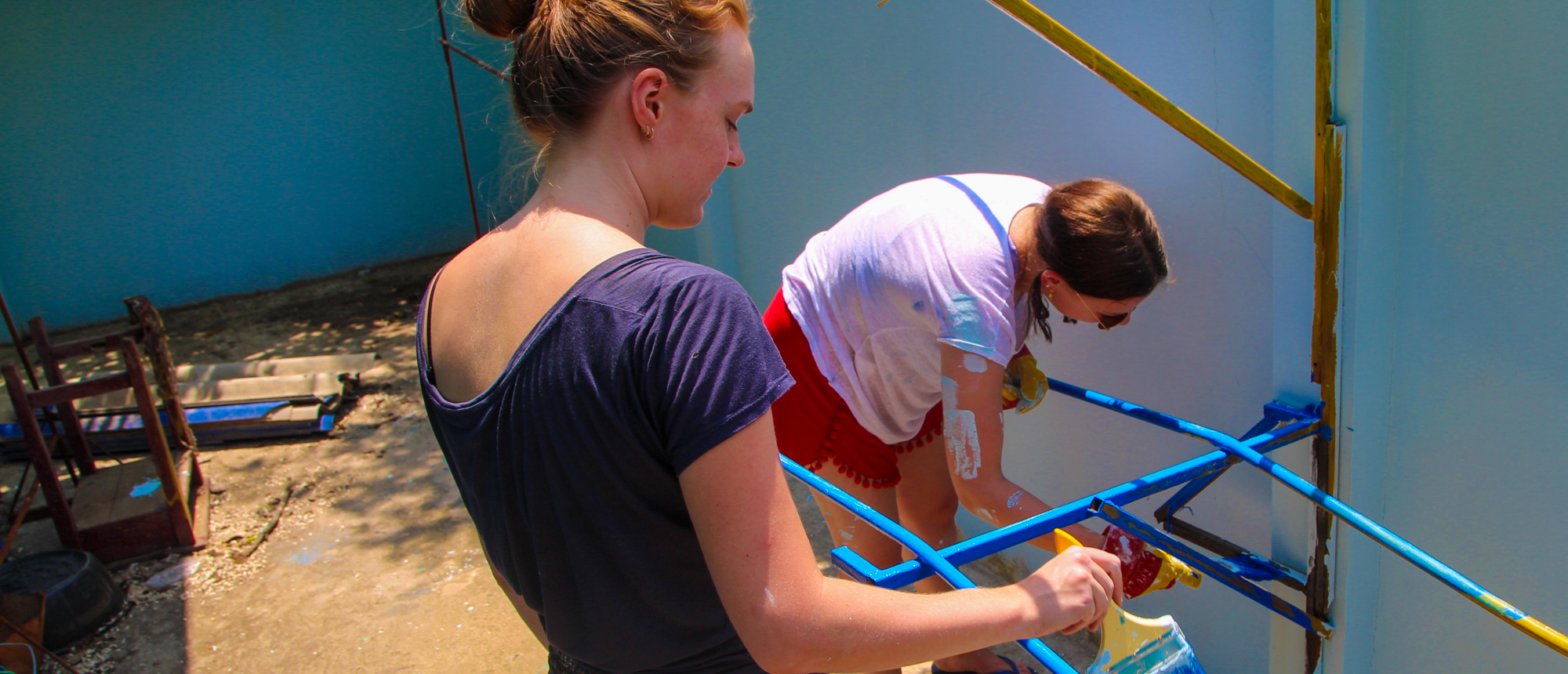 Two women volunteers painting blue metal bars and a light blue wall outdoors.