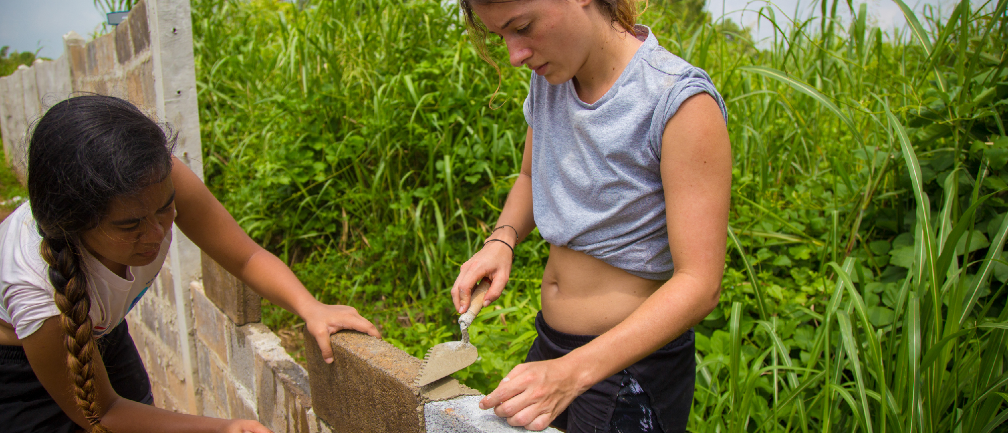 Two women volunteer, building a concrete block wall outdoors in a green, tropical setting.