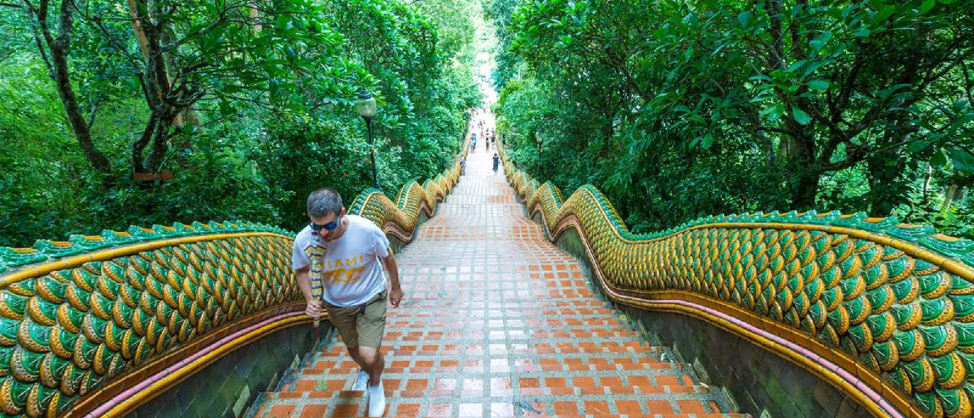 A traveler climbs the long, ornate Naga staircase at Doi Suthep temple, surrounded by lush trees.
