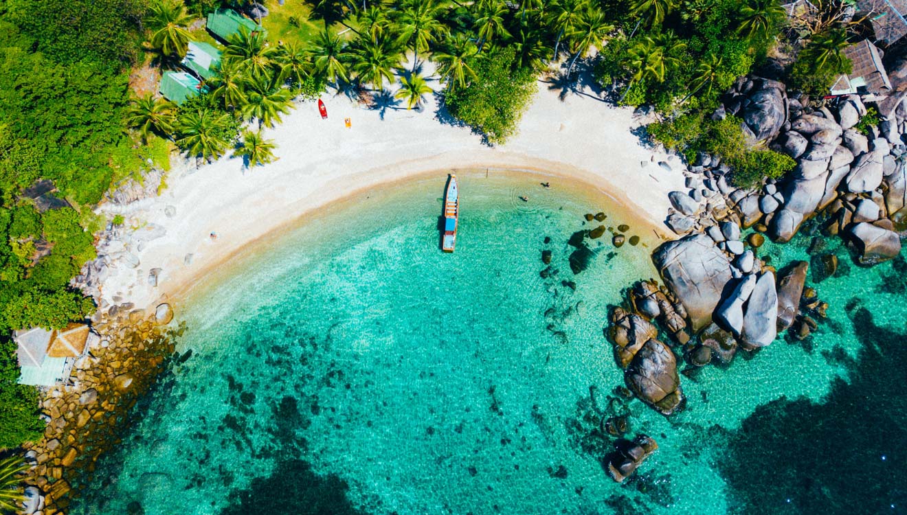 Aerial view of a longtail boat on a white sand beach with turquoise water in Koh Phangan, Thailand.