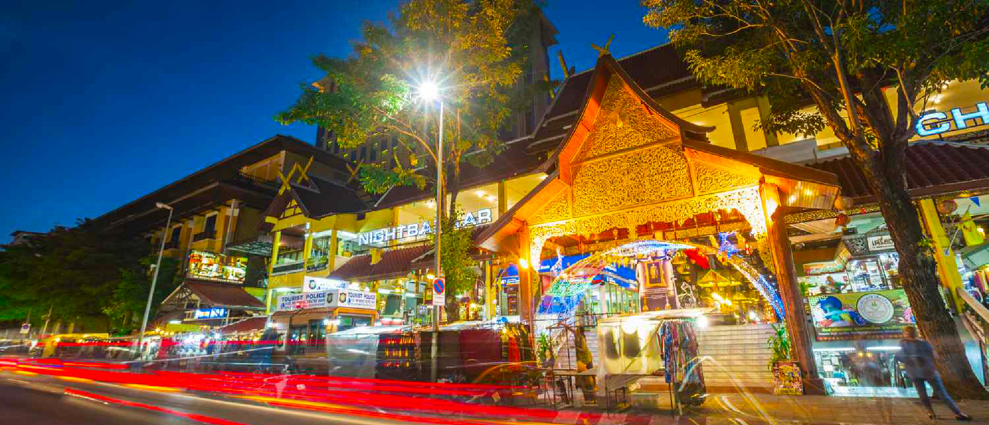 Vibrant night scene in Chiang Mai with traditional Thai buildings, bright streetlights, and red light trails.