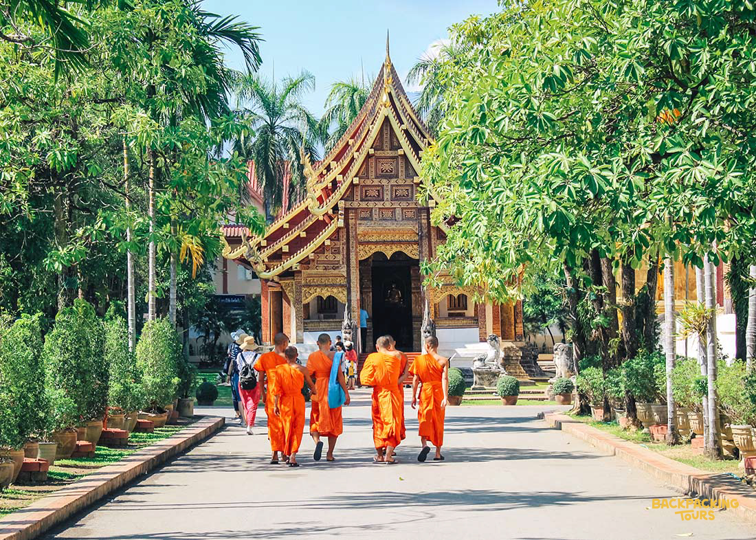 Monks in orange robes walk towards a beautiful golden temple surrounded by green trees in Thailand.