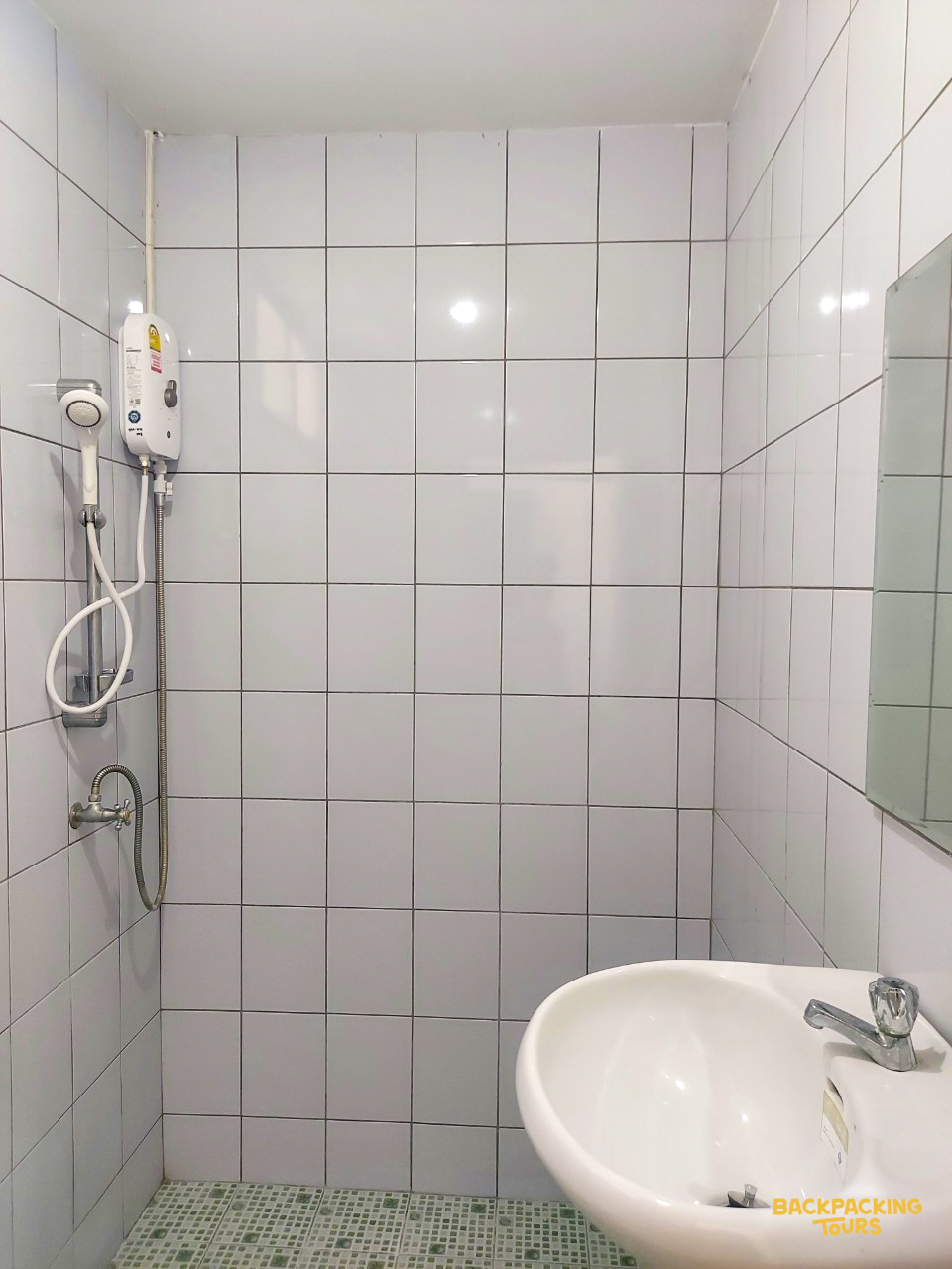 Functional bathroom with white tiles, an electric shower heater, a sink, and a mirrored wall.