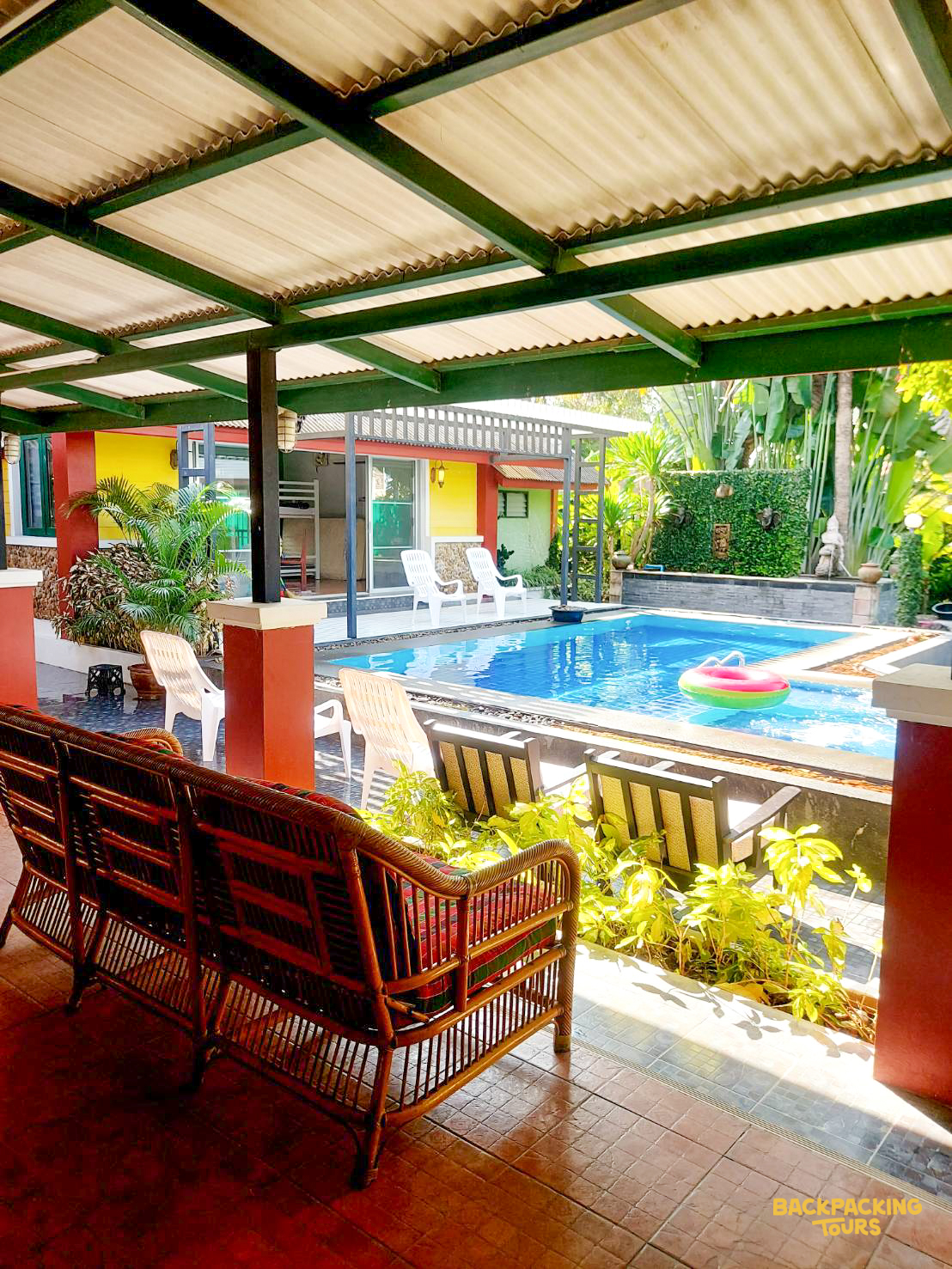 A comfortable rattan sofa on a covered patio overlooking a vibrant swimming pool and tropical buildings in Thailand.