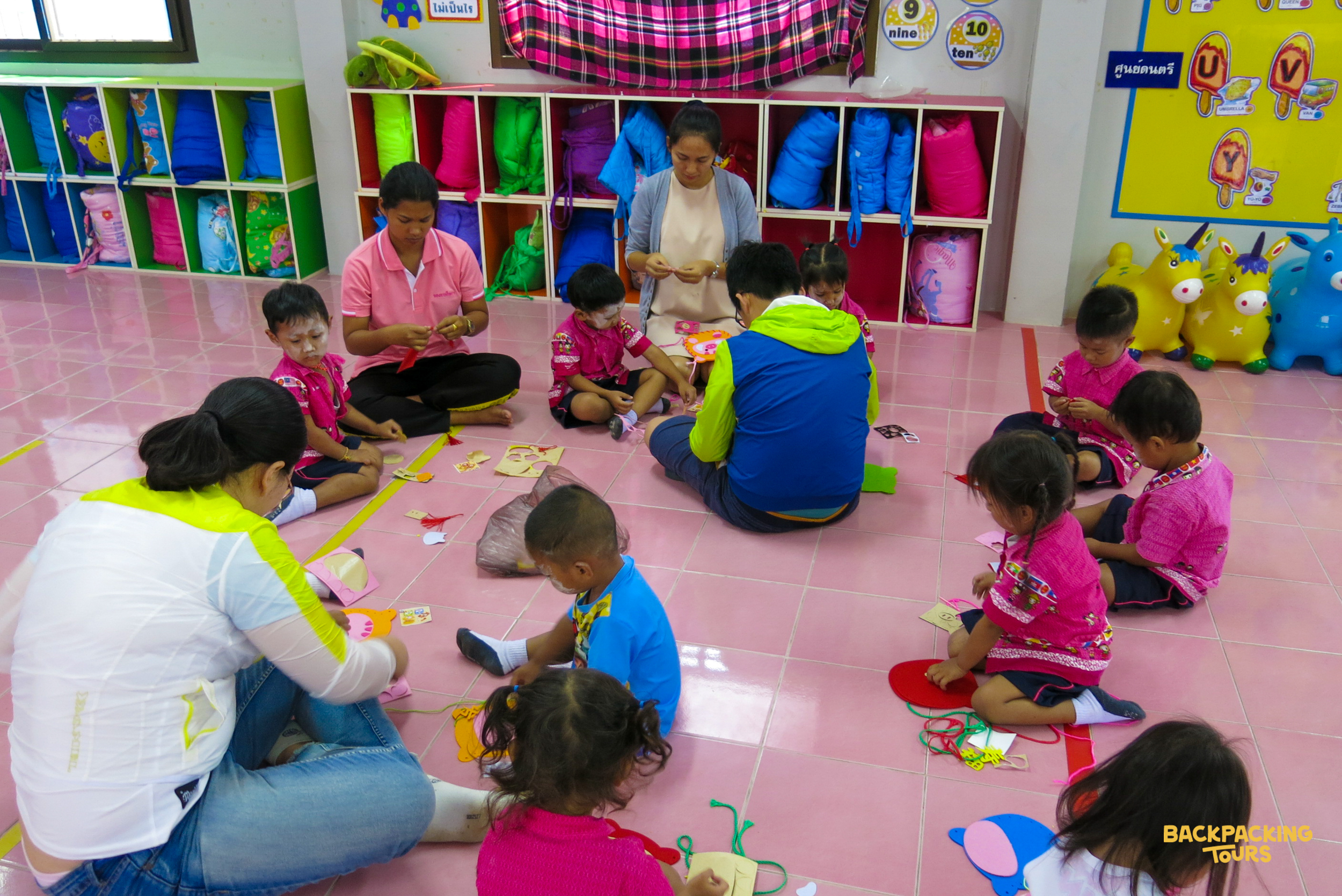 Young children and volunteers sit on the floor, making colorful crafts in a bright Thai kindergarten classroom.
