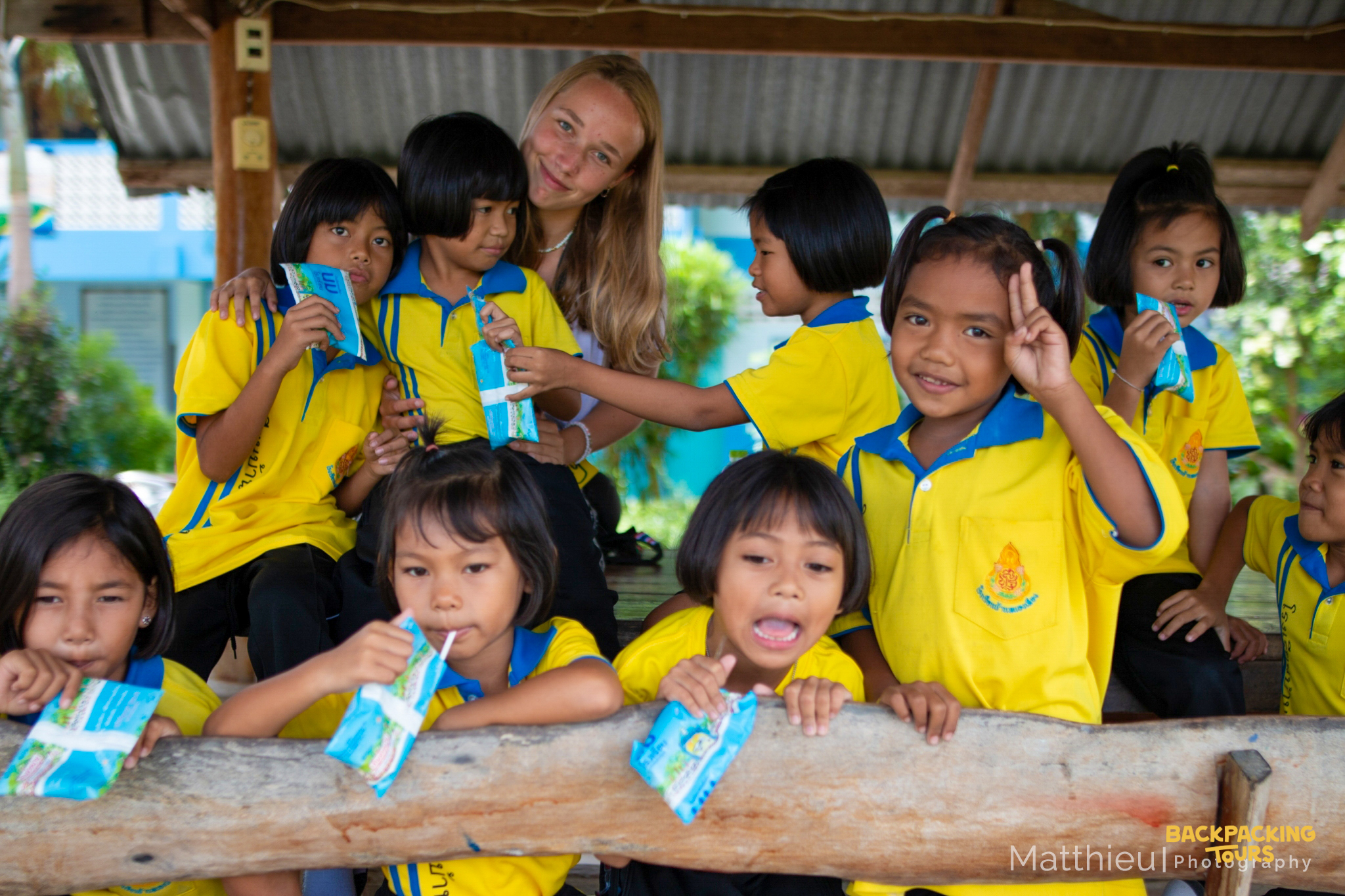 A volunteer smiles with happy Thai kindergarten children drinking juice under a shelter.