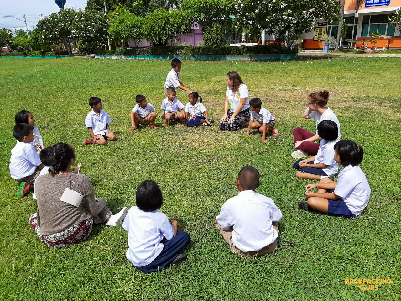 Volunteers and children sit in a circle on green grass, engaging in a fun activity in Thailand.