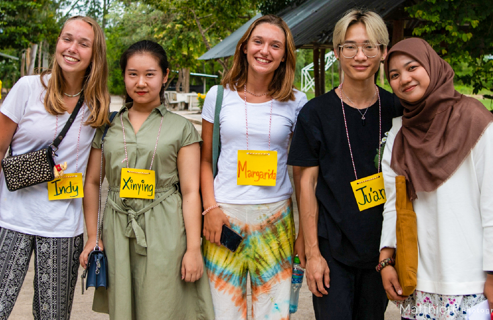 Five diverse young volunteers smiling together outdoors in a rural setting in Thailand.